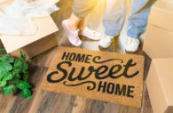 Man and Woman Standing Near Home Sweet Home Welcome Mat, Moving Boxes and Plant.